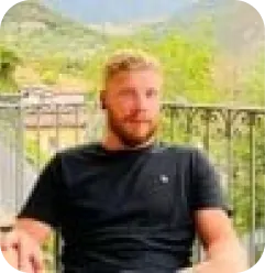 A man with short blond hair and a beard sits outdoors on a patio, wearing a black t-shirt. Behind him is a metal railing and a green, mountainous landscape.