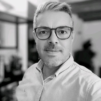 A man with short, light hair and glasses looks at the camera, wearing a light-colored button-up shirt. The background is blurred, with faint outlines of furniture and a window. The image is in black and white.