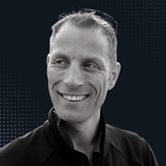 A man with short light hair, wearing a zipped-up jacket and sunglasses on his head, smiles while looking to the side. The background is dark with a subtle dotted pattern.