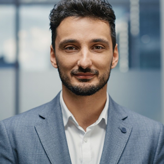 A man with short dark hair and a trimmed beard is wearing a light blue suit jacket and white shirt, smiling slightly, standing indoors with blurred office windows in the background.