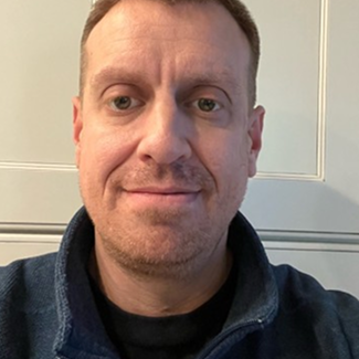 A man with short light brown hair and fair skin is wearing a navy blue fleece jacket and smiling slightly while looking at the camera. He is standing indoors in front of a light-colored wall.
