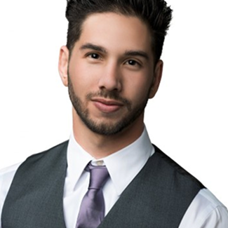A young man with dark hair and a trimmed beard, wearing a white shirt, light purple tie, and dark vest, poses against a plain white background, looking confidently at the camera.