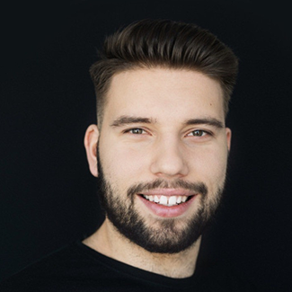 A young man with short dark hair and a full beard is smiling at the camera against a plain black background. He is wearing a black shirt.