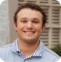 A young man with curly brown hair smiles at the camera. He is wearing a light blue collared shirt with a striped undershirt. The background shows a building with windows and light-colored bricks.
