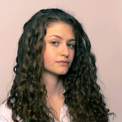 A young woman with long, curly brown hair and light eyes looks at the camera with a neutral expression. She is wearing a light-colored shirt and is posed against a plain, light background.
