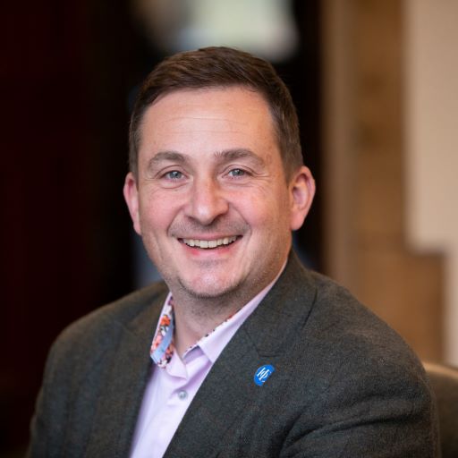 A smiling man in a gray blazer and light-colored shirt with a floral collar detail, wearing a blue pin, sits indoors with a blurred background.